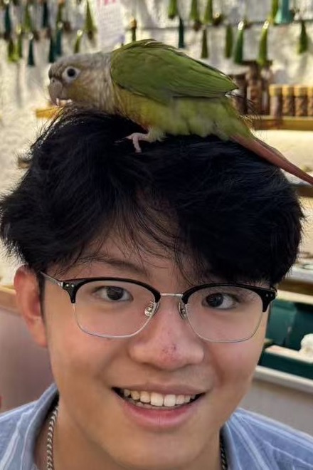 Charles Zheng, smiling, with a green parakeet perched on his head